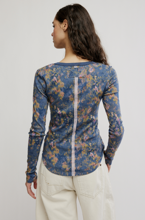 A woman shown from the back wearing a fitted long-sleeve top with a blue floral print and a vertical white zipper detail, paired with cream pants, against a plain light gray studio background.