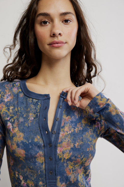 A close-up of a woman wearing a long-sleeve button-front top with a blue base and a multicolor floral print in pink, yellow, and orange, against a light gray background.