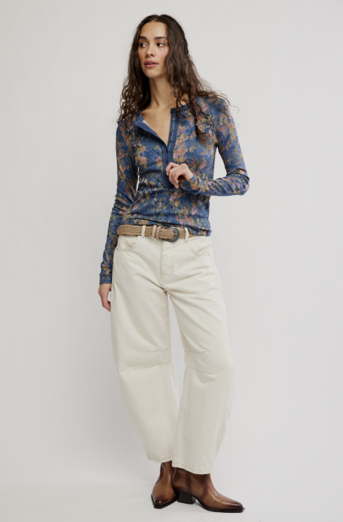 A woman modeling a blue and gold abstract patterned zip-up shirt, cream wide-leg pants, a woven belt, and brown leather boots against a light gray studio background.
