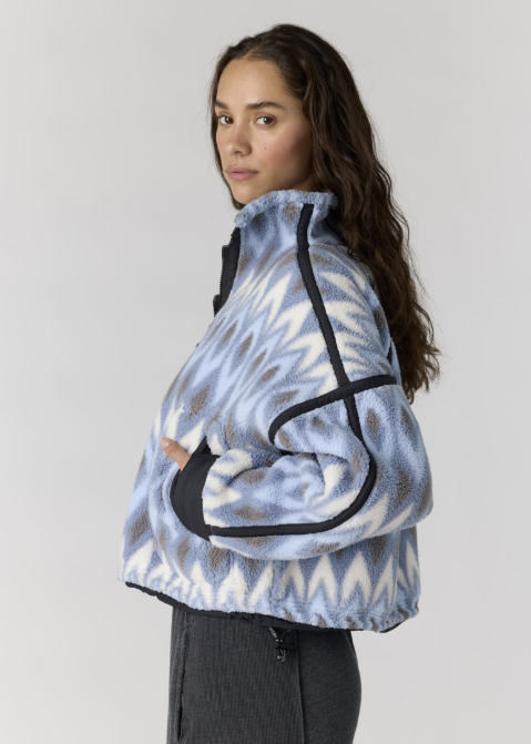A woman wearing a cropped fleece jacket with a light blue, white, and gray chevron pattern and black contrast piping, shown from a side profile against a light gray studio background.