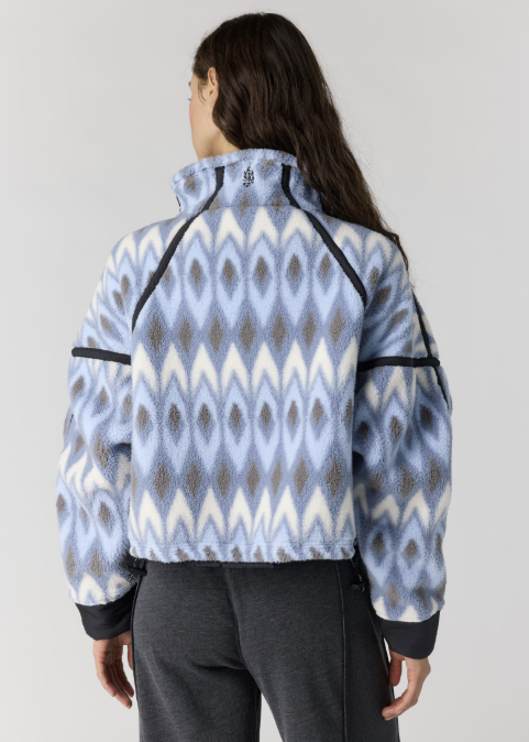 A woman shown from the back wearing a cropped fleece jacket with a light blue, white, and gray ikat pattern, paired with dark gray sweatpants, against a plain light gray studio background.