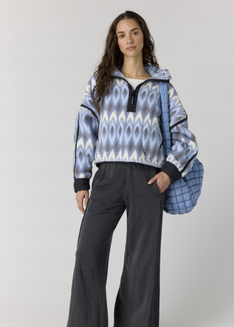 A model stands against a light gray studio background wearing a blue, white, and gray patterned half-zip pullover, dark gray wide-leg pants, and carrying a blue quilted shoulder bag.