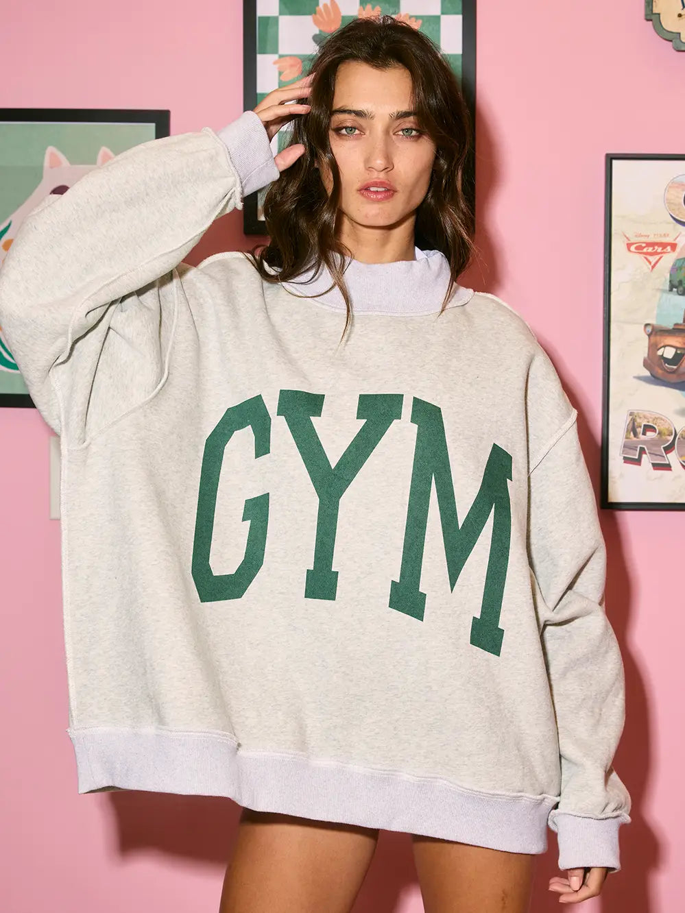 Woman wearing a gray sweatshirt with 'GYM' printed in green letters against a pink wall with framed pictures.