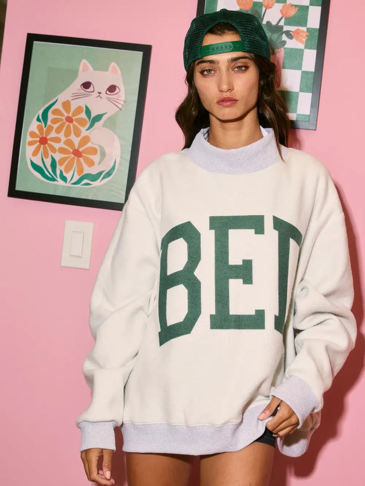 Person wearing a white sweatshirt with green text in a room with pink walls and framed artwork.