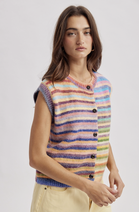 A woman wearing a sleeveless, button-front knit vest with horizontal multicolor stripes in shades of blue, purple, pink, orange, yellow, and green, paired with light beige pants, against a plain light gray studio background.