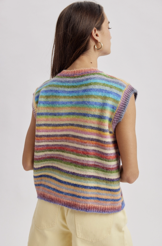 A woman shown from the back wearing a sleeveless, ribbed knit sweater with horizontal multicolor stripes in shades of blue, green, pink, orange, yellow, and purple, paired with light beige pants against a light gray studio background.