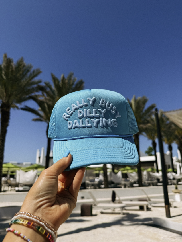 Blue cap with text held by a hand against a palm tree-lined background