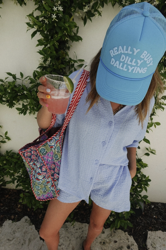 Person wearing a blue cap with text, holding a drink and colorful bag, standing against a green leafy background.