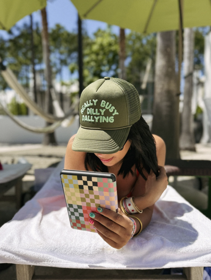 Person wearing a green cap with text, sitting under an umbrella at a beach or poolside.