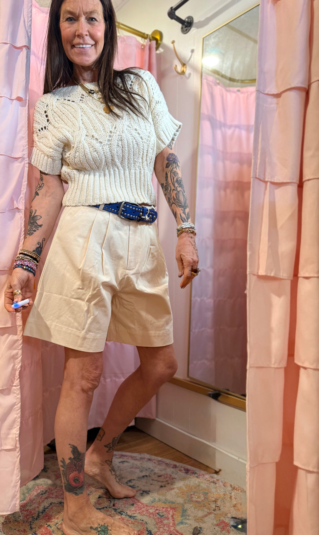 Woman standing in a room with pink curtains and a mirror, wearing a white knit top and beige shorts.