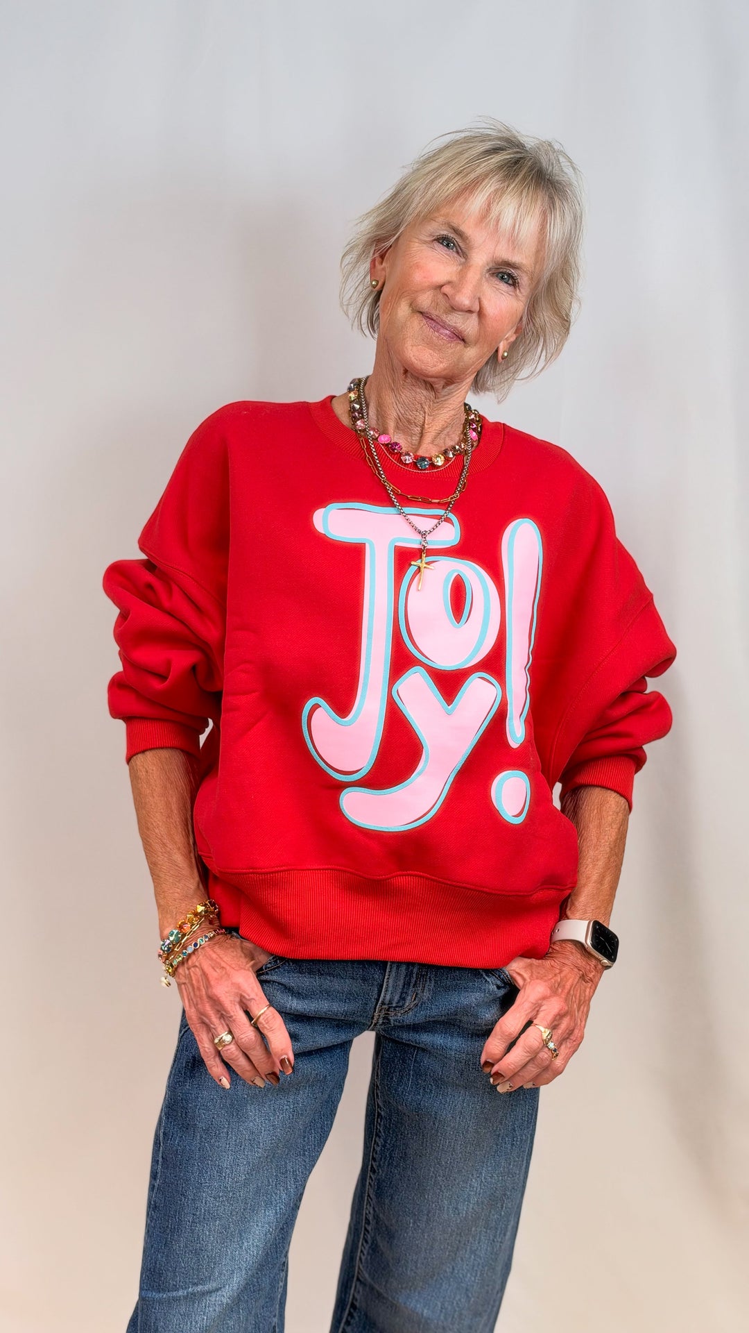 Woman wearing red sweatshirt with 'JOY!' in pink and blue on plain background