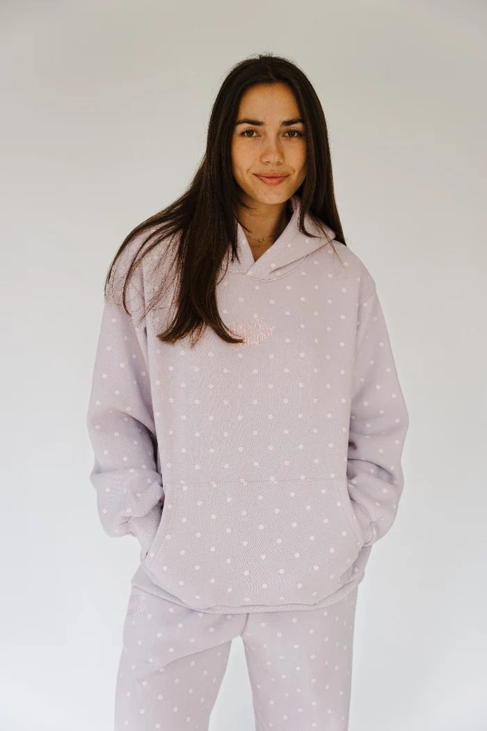 A woman modeling a light lavender pajama set with a white polka dot pattern, consisting of a hooded sweatshirt and matching pants, against a plain light gray background.