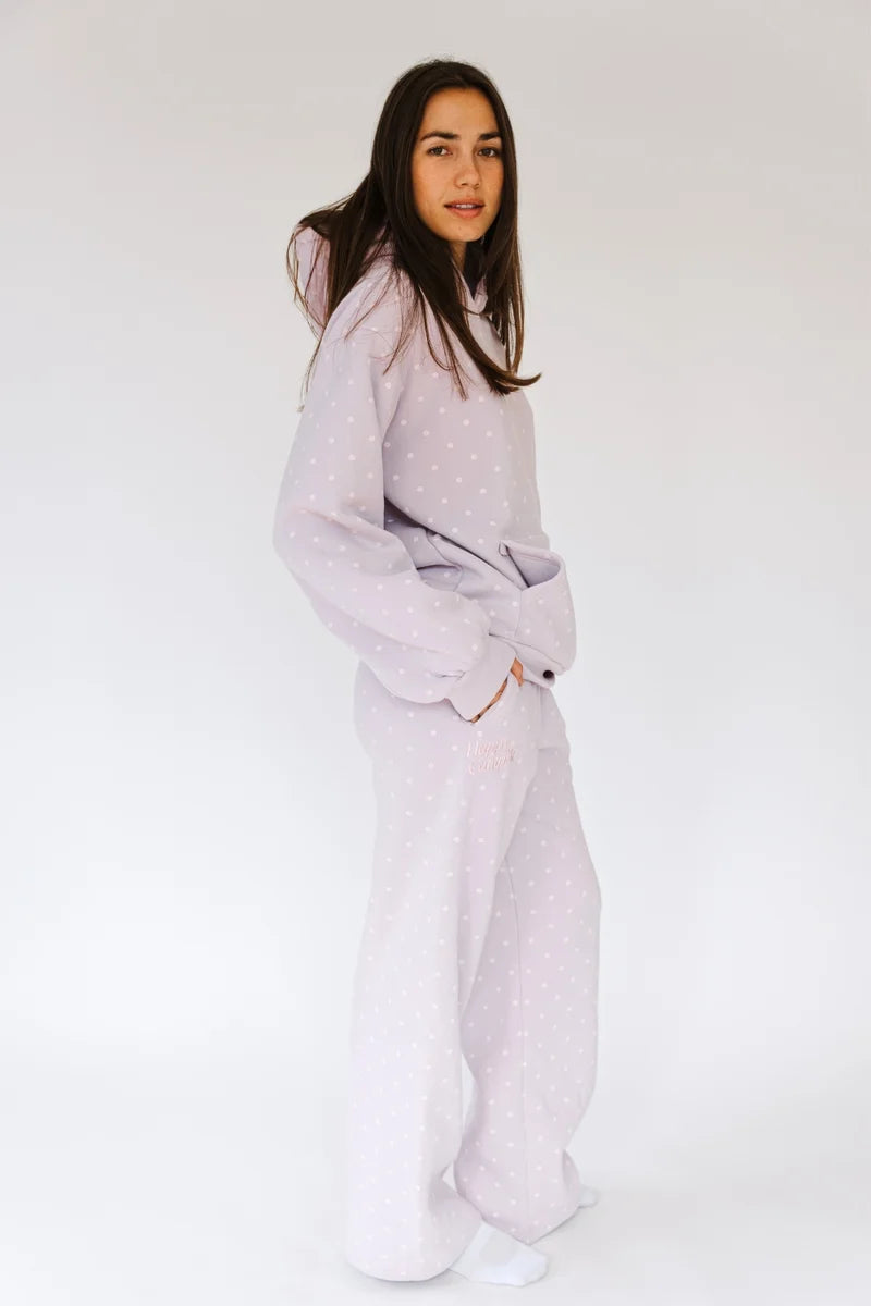 A woman modeling a light lavender pajama set with a subtle white polka dot pattern, consisting of a long-sleeve top and matching wide-leg pants, against a plain white studio background.