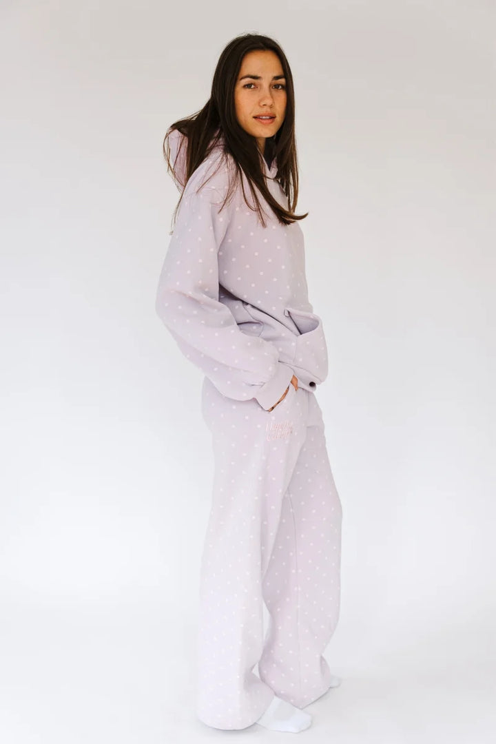 A woman modeling a light lavender pajama set with a subtle white polka dot pattern, consisting of a long-sleeve top and matching wide-leg pants, against a plain white studio background.