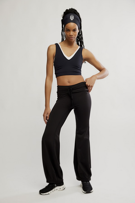 A model wearing a matching black activewear set consisting of a V-neck sports bra with a white trim, black drawstring sweatpants, a black headband with a white logo, and black sneakers, posed against a light gray studio background.