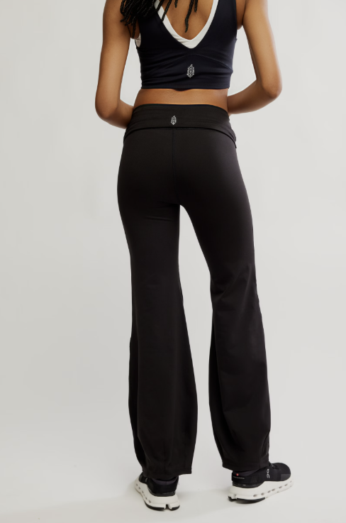 Rear view of a model wearing black high-waisted flared leggings or yoga pants with a small metallic logo at the center of the waistband, paired with a dark sports bra and black sneakers, against a light gray studio background.