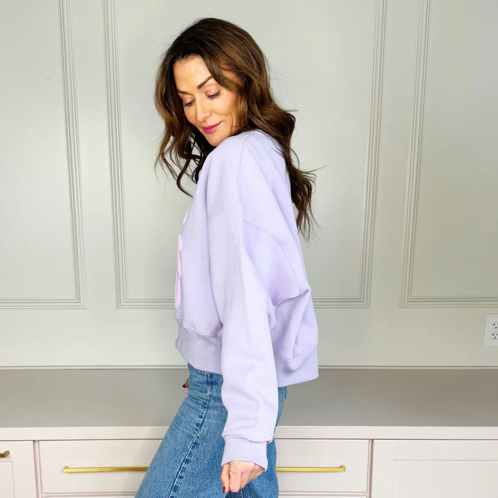 Woman wearing a light purple sweatshirt and blue jeans in a kitchen setting.