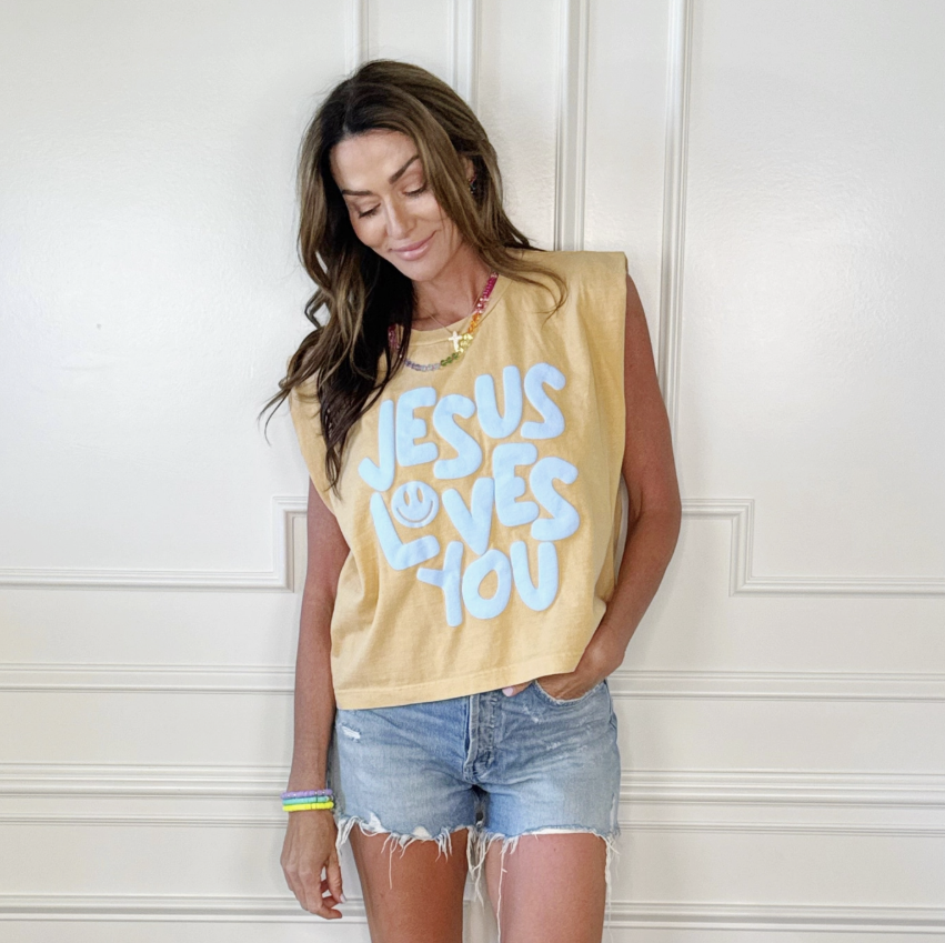 Woman wearing a yellow tank top with 'Jesus Loves You' text against a white wall.