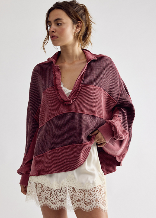 A woman models a loose, oversized knit top with horizontal stripes in dusty rose and dark plum, featuring a ruffled collar and long balloon sleeves, paired with white lace-trimmed shorts against a light gray studio background.