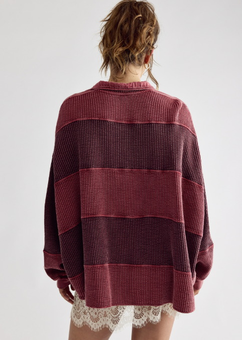 A woman viewed from behind wearing an oversized ribbed knit sweater with wide horizontal stripes in dusty rose and deep burgundy, paired with white lace shorts against a light gray studio background.