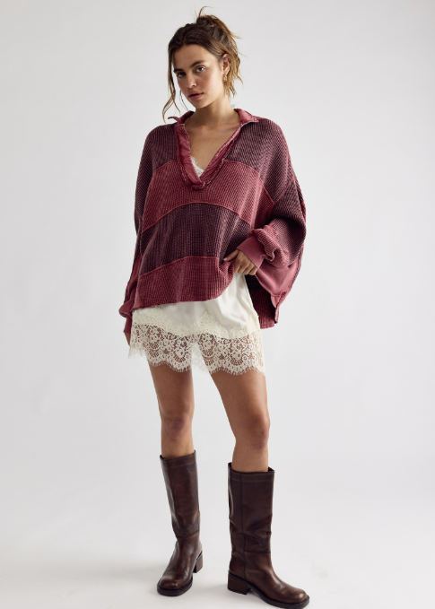 A model stands against a light gray studio background wearing a loose, oversized knit poncho-style top in dark burgundy and maroon tones, layered over a white lace-trimmed skirt or dress, with dark brown knee-high boots.