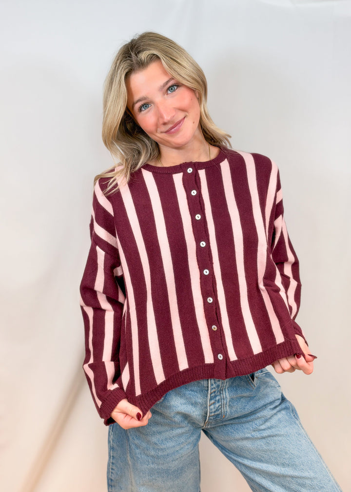 Woman wearing burgundy and pink stripe cardigan with jeans on plain background