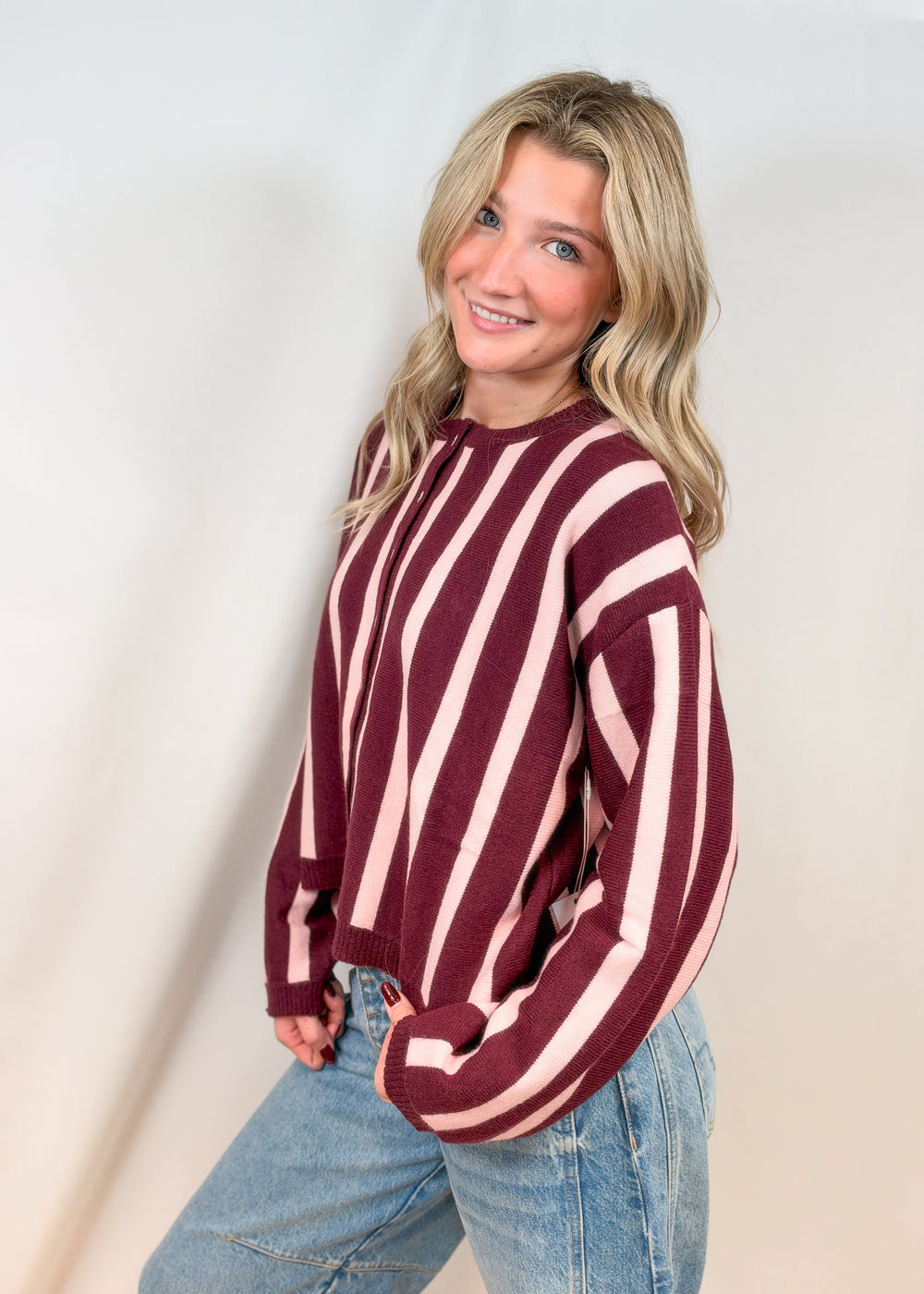 Woman shown from side view wearing burgundy and pink stripe cardigan with jeans on plain background