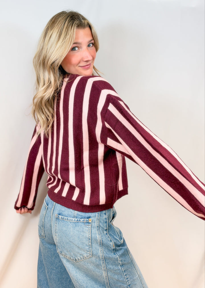 Woman shown from back view wearing burgundy and pink stripe cardigan with jeans on plain background