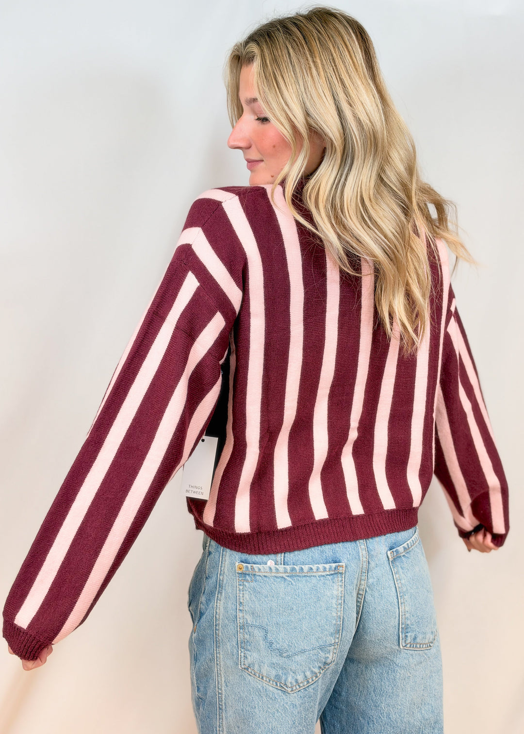 Woman shown from back view wearing burgundy and pink cardigan with jeans on plain background
