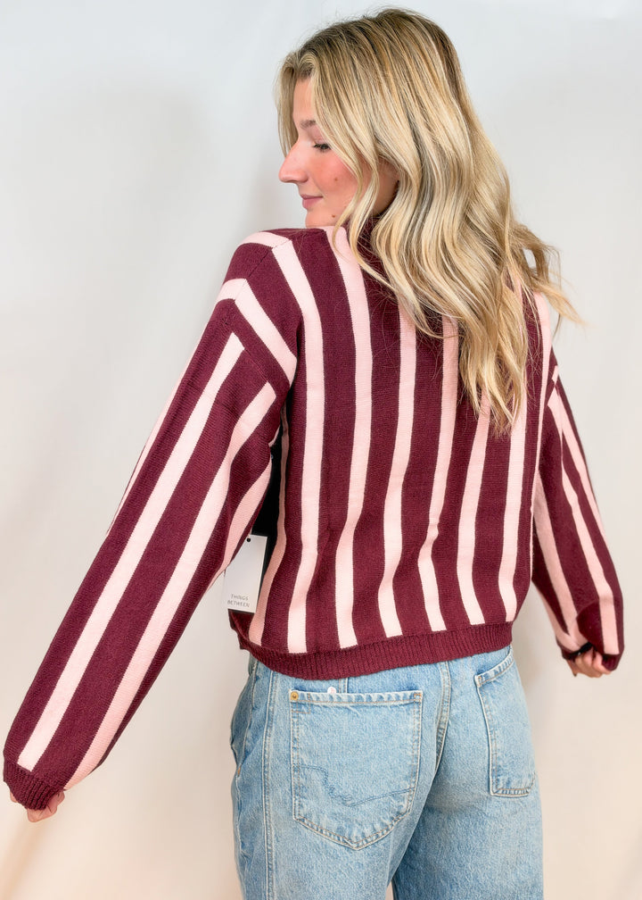 Woman shown from back view wearing burgundy and pink cardigan with jeans on plain background
