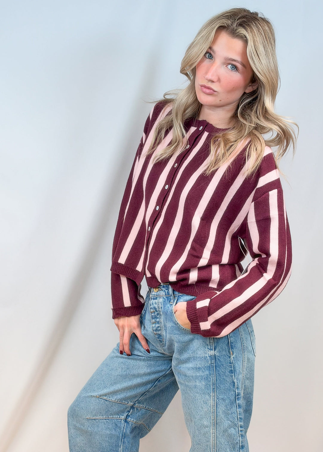 Woman wearing burgundy and pink stripe cardigan with jeans on plain background