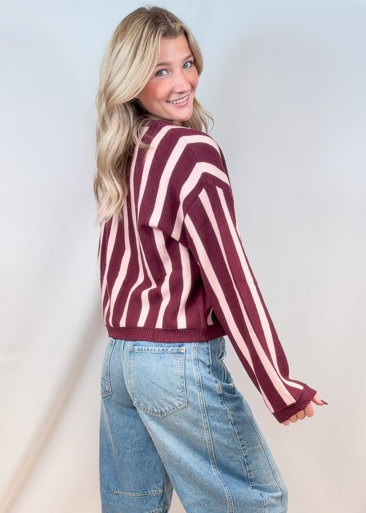 Woman shown from back wearing burgundy and pink stripe cardigan with jeans on plain background