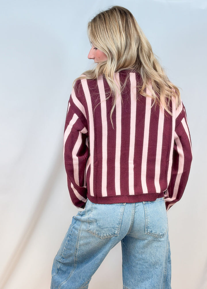 Woman shown from back wearing burgundy and pink stripe cardigan with jeans on plain background