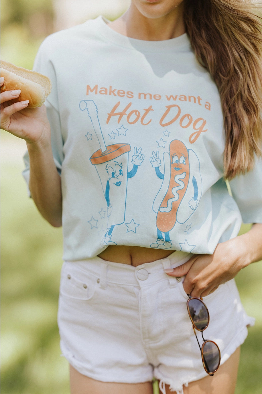 Woman wearing graphic tee with hot dog and drink with shite shorts standing outdoors