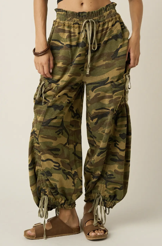 A person wearing loose camouflage harem-style pants with drawstring waist and ankle ties, shown from the waist down against a plain light beige studio background.