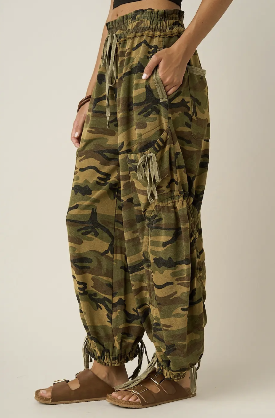 A person wearing loose camouflage harem-style pants with drawstrings, side pockets, and fringe details, paired with brown sandals against a plain light beige studio background.