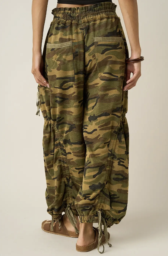A person shown from the back wearing loose camouflage harem-style pants with drawstring hems and side cargo pockets, paired with tan flat sandals against a plain light beige studio background.