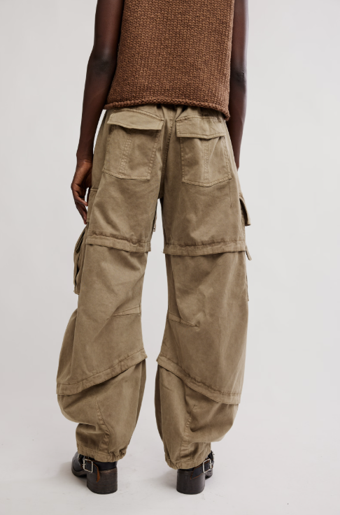 A model shown from the back wearing baggy olive-brown cargo pants with multiple flap pockets, a brown knit sleeveless top, and black shoes against a plain light gray studio background.