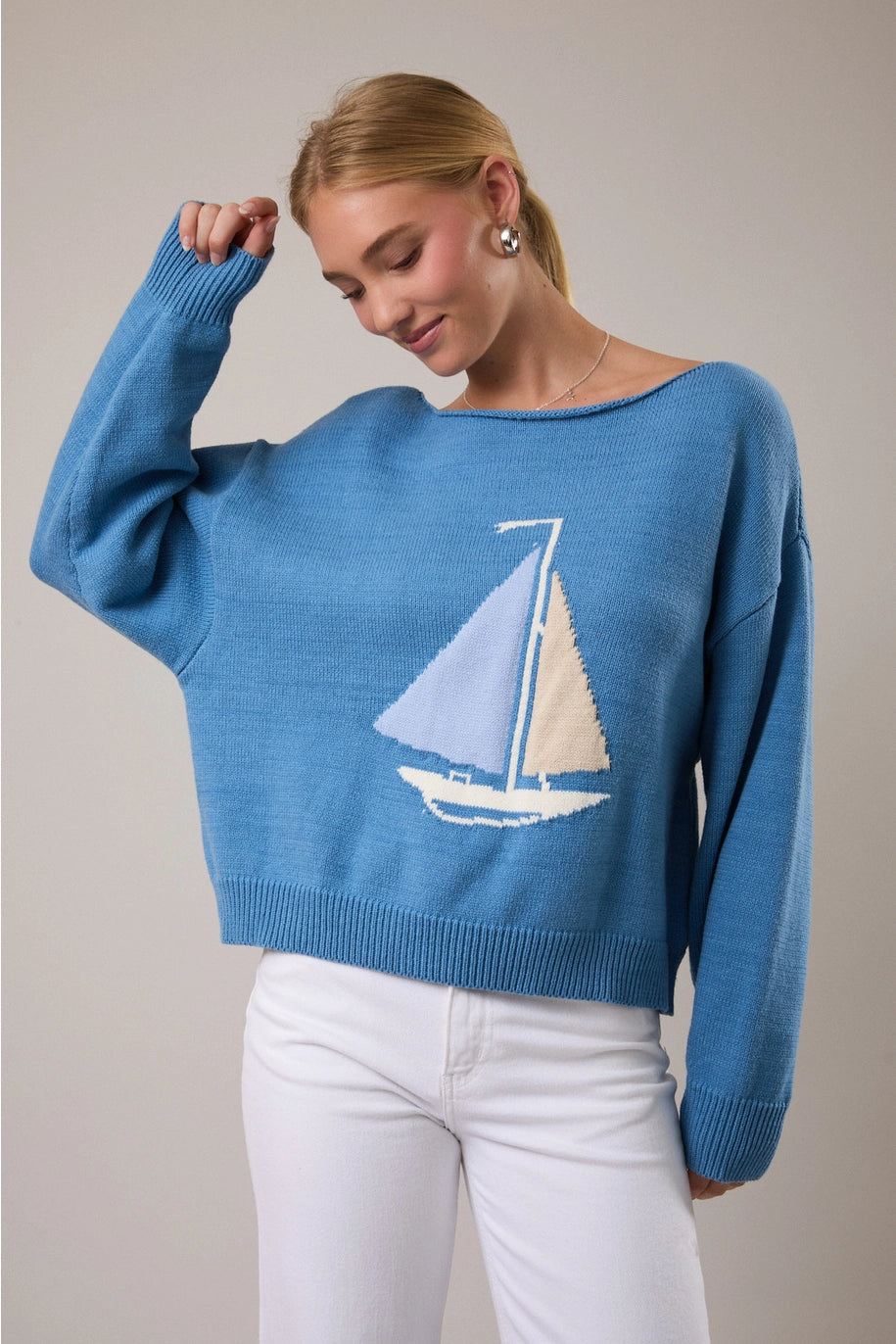 Woman wearing blue sweater with boat on front with white pants on plain background