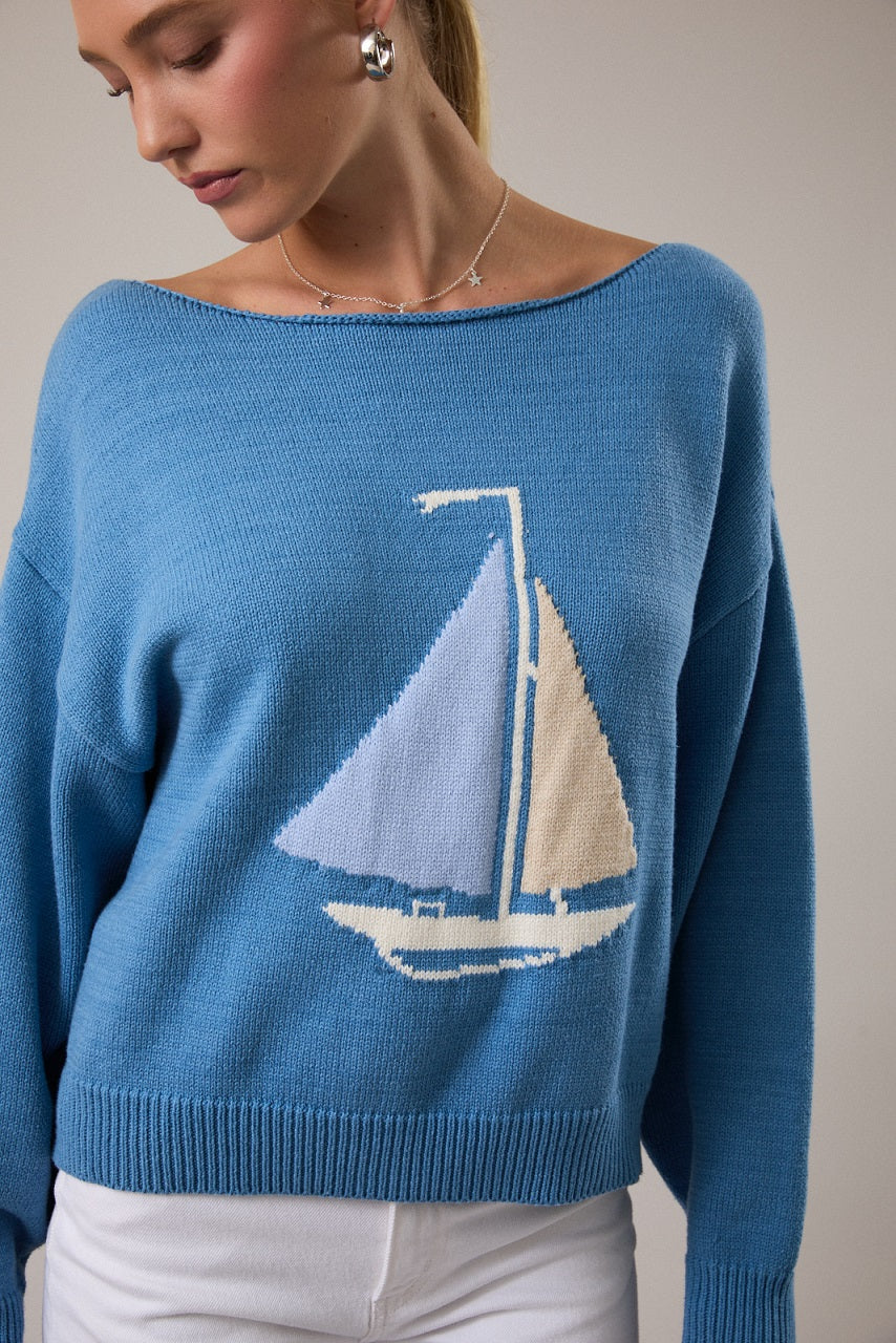 Woman wearing blue sweater with boat detail and white pants on plain background