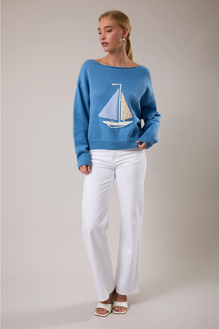 Woman wearing blue sweater with boat detail and white pants on plain background