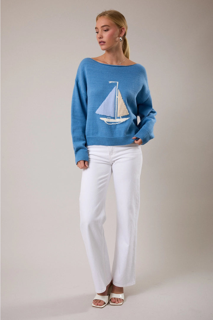 Woman wearing blue sweater with boat detail and white pants on plain background
