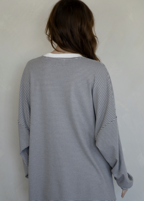 A person viewed from the back wearing an oversized long-sleeve top with thin horizontal gray and white stripes, layered over a white crewneck shirt, against a plain light gray wall.