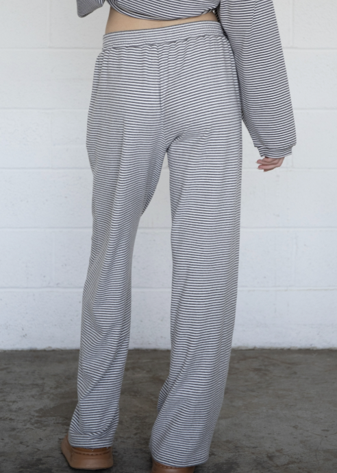 A person shown from the back wearing loose, wide-leg pants and a matching long-sleeve crop top with thin horizontal gray and white stripes, standing against a white painted brick wall on a concrete floor.
