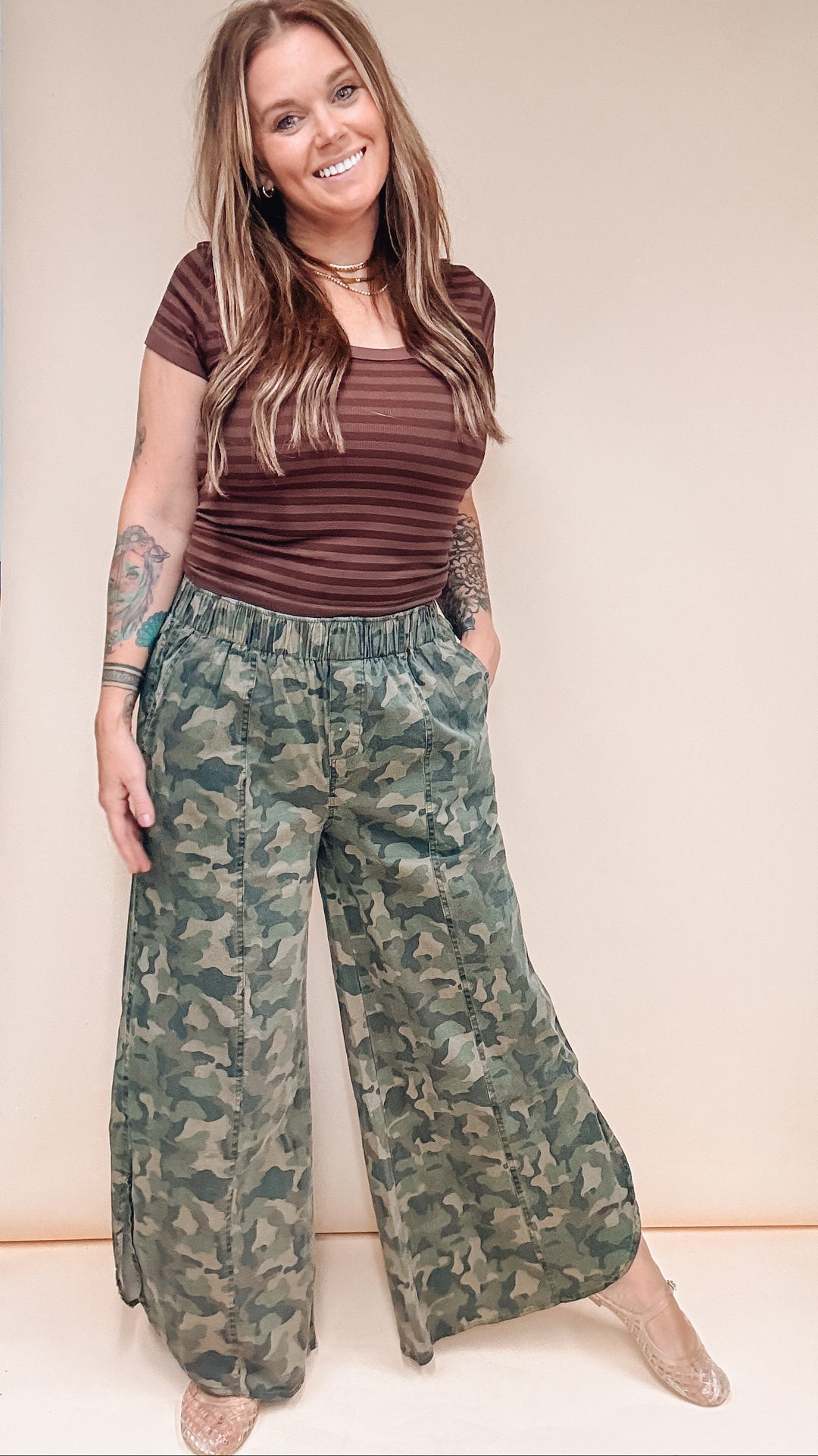 A woman modeling a pair of wide-leg camouflage pants and a brown striped t-shirt against a plain beige studio background.