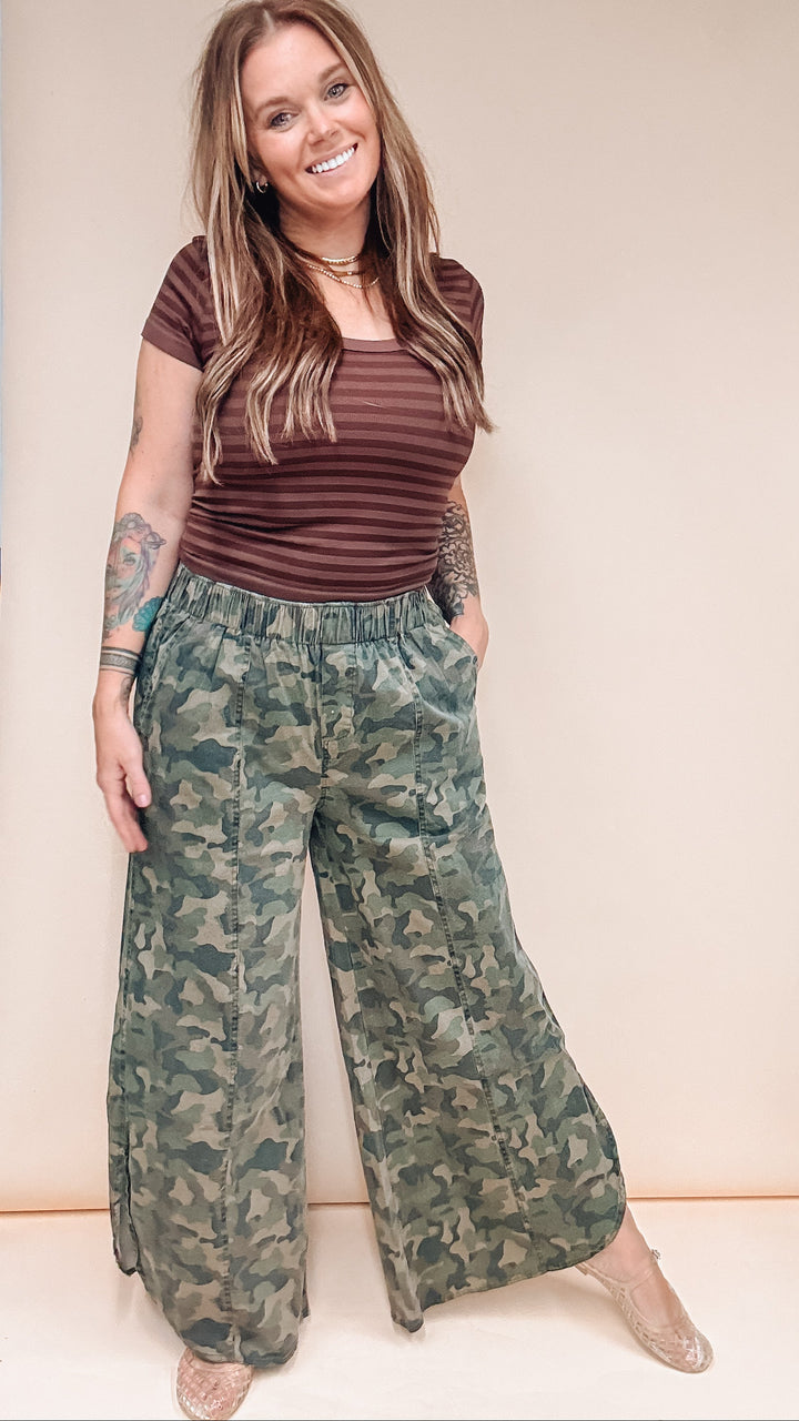 A woman modeling a pair of wide-leg camouflage pants and a brown striped t-shirt against a plain beige studio background.
