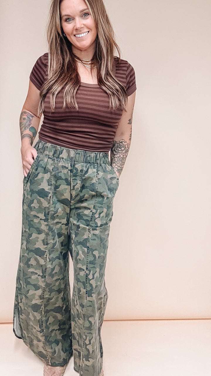 A woman modeling a brown and burgundy striped short-sleeve top with green camouflage wide-leg pants, standing against a solid beige studio background.