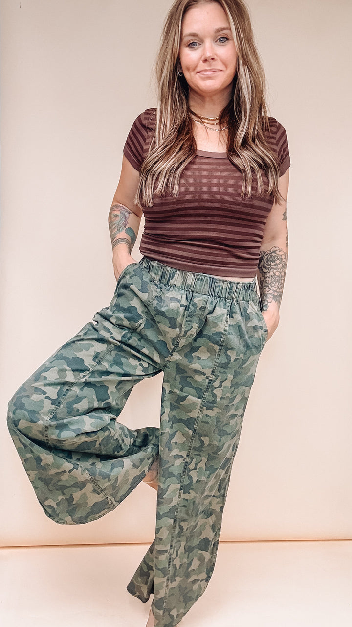 A woman modeling a pair of wide-leg camouflage pants and a brown striped t-shirt against a solid beige studio background.
