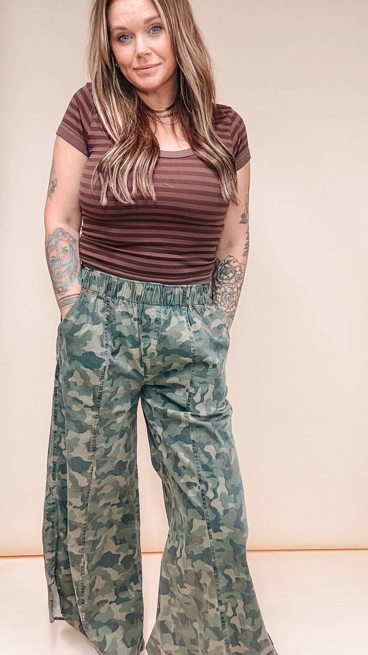 A woman modeling a brown and dark brown striped short-sleeve t-shirt and green camouflage wide-leg pants, standing against a plain beige studio background.