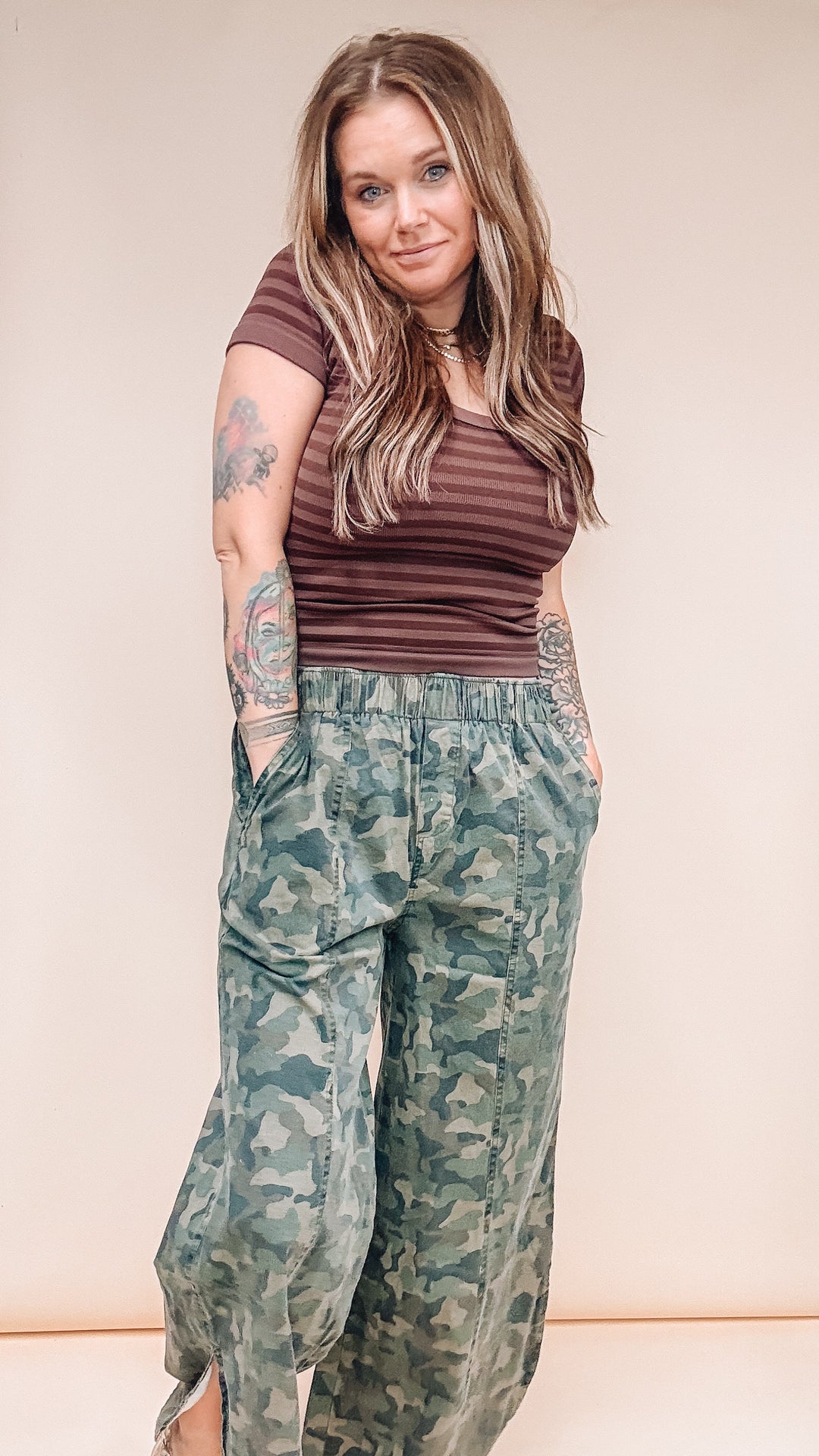 A woman modeling a brown striped short-sleeve top and green camouflage wide-leg pants against a plain beige studio background.